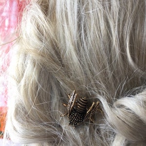 Puede incluir: Una pinza para el pelo de insecto dorada con un cuerpo texturizado y diamantes de imitaci&oacute;n transparentes. La pinza est&aacute; en cabello rubio.