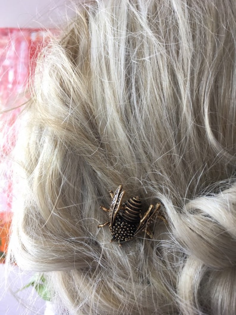 Puede incluir: Una pinza para el pelo de insecto dorada con un cuerpo texturizado y diamantes de imitaci&oacute;n transparentes. La pinza est&aacute; en cabello rubio.