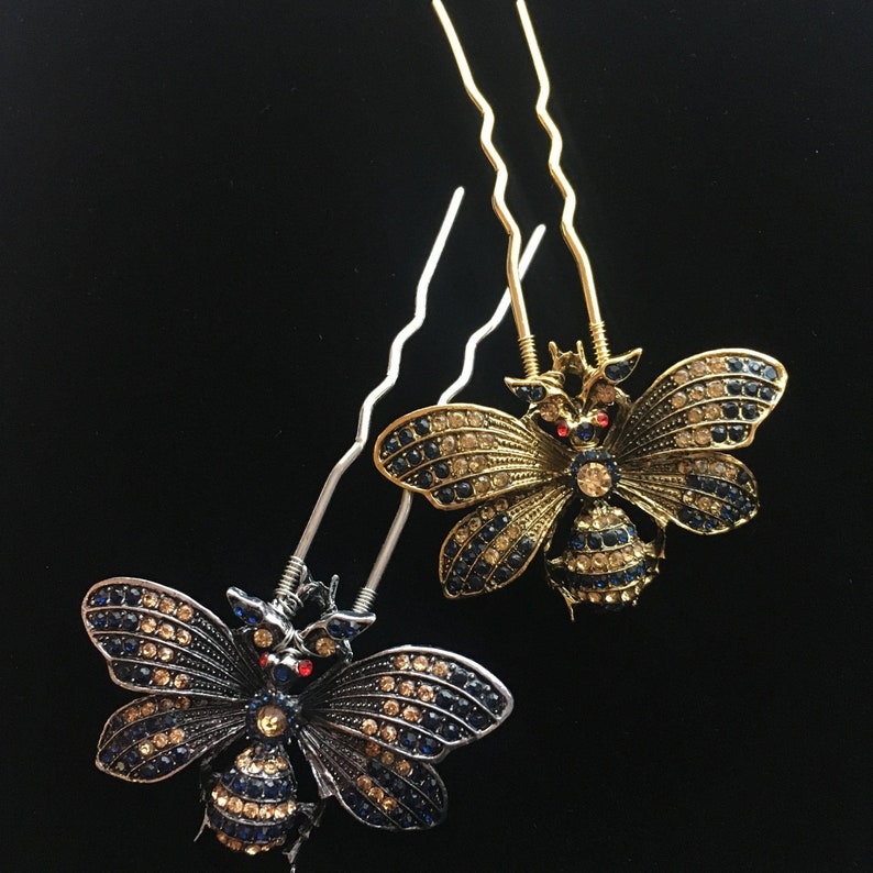 Puede incluir: Dos horquillas doradas con dise&ntilde;os de mariposas adornadas con joyas. Una mariposa es azul y negra con detalles rojos, la otra es dorada con detalles azules y transparentes.