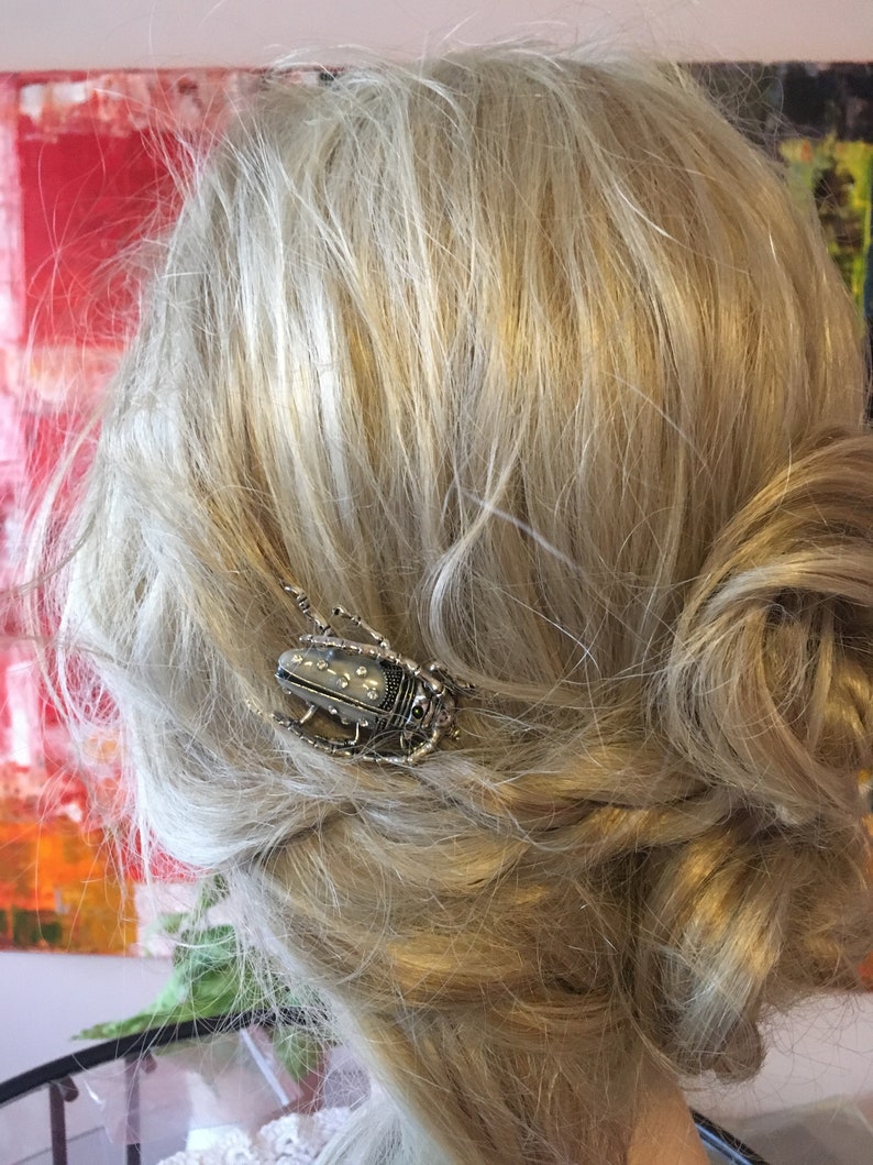Puede incluir: Una pinza para el pelo de plata en forma de escarabajo con un dise&ntilde;o blanco y negro. La pinza est&aacute; en el pelo rubio de una mujer.