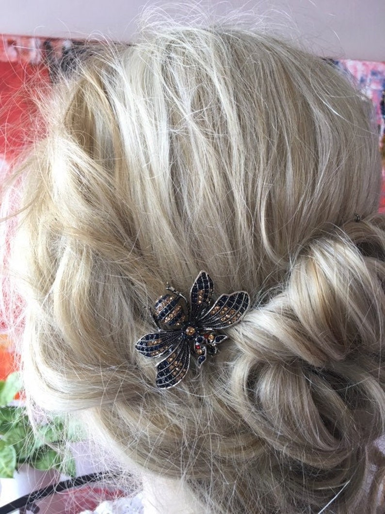 Puede incluir: Una pinza para el pelo en forma de abeja negra y dorada con pedrer&iacute;a. La pinza est&aacute; en el pelo de una persona con el pelo rubio.