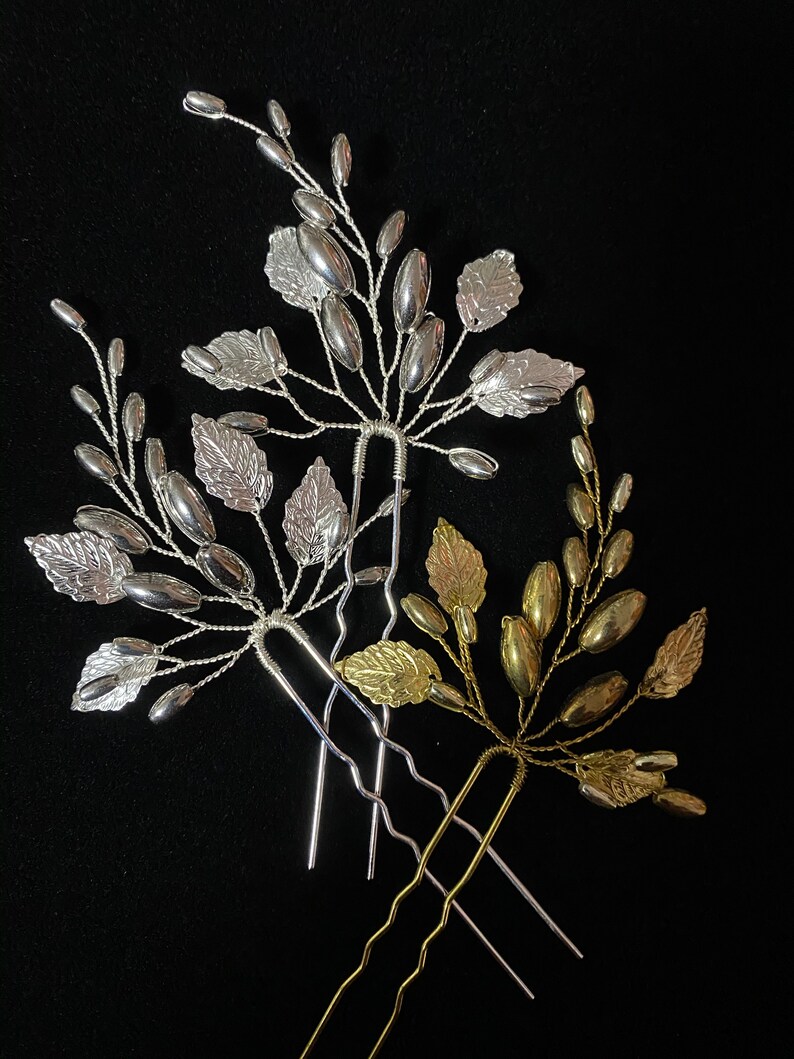 May include: Three silver and gold hair pins with leaf and bead accents. The pins are arranged in a fan shape on a black background.