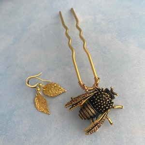 Puede incluir: Una pinza para el pelo de abeja dorada con pedrer&iacute;a y un par de pendientes de hoja dorada. La abeja es negra con detalles dorados y tiene un cuerpo texturizado.