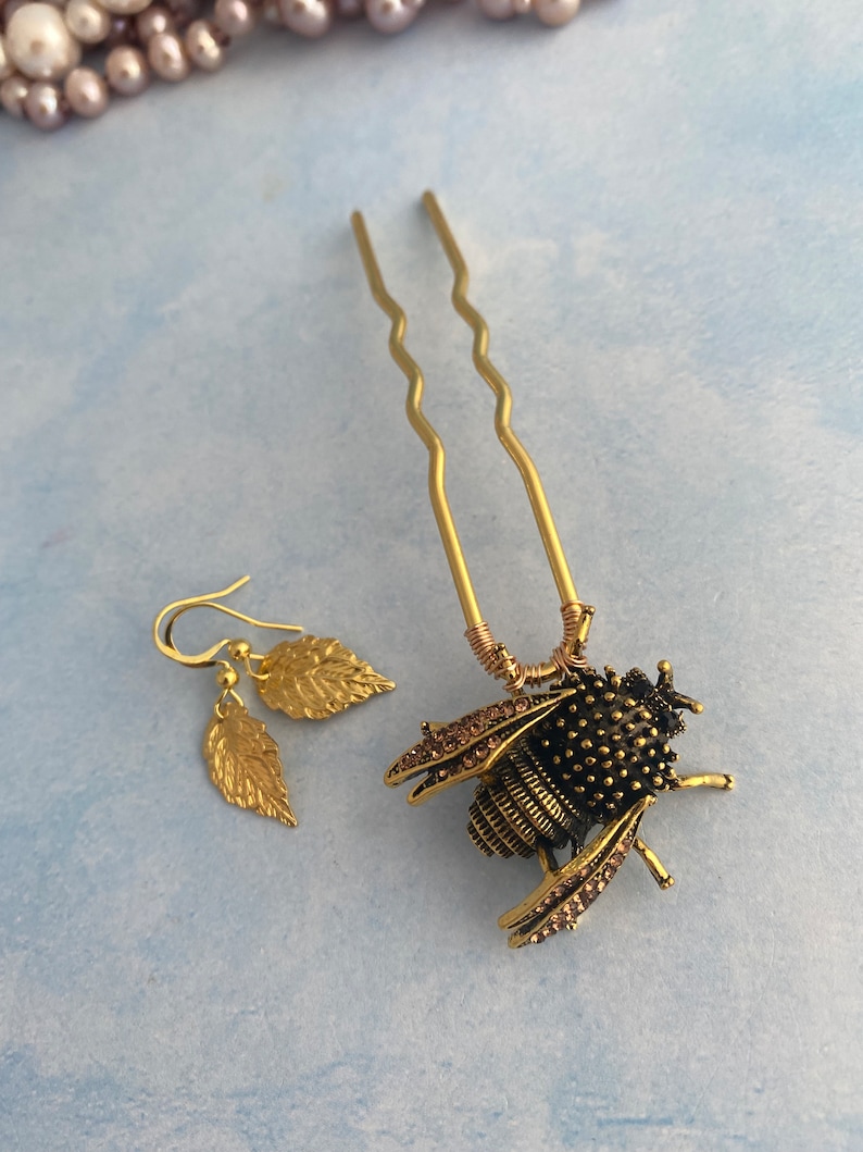 Puede incluir: Una pinza para el pelo de abeja dorada con pedrer&iacute;a y un par de pendientes de hoja dorada. La abeja es negra con detalles dorados y tiene un cuerpo texturizado.