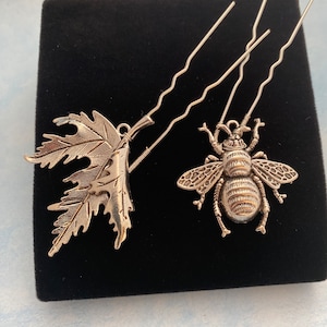 May include: Two silver hair clips, one shaped like a leaf and the other like a bee. The bee has a textured body and wings.