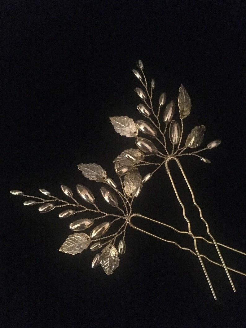 May include: Two gold hair pins with leaf and bead embellishments. The pins are designed to look like branches with leaves and small, oval-shaped beads.