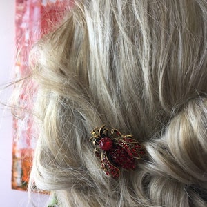 Puede incluir: Una pinza para el pelo roja y dorada con joyas en forma de insecto estilizado. La pinza est&aacute; en el pelo rubio de una mujer.