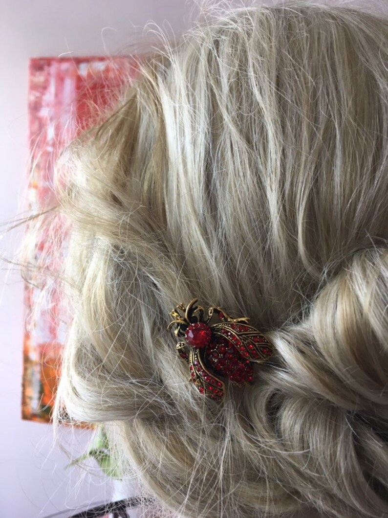 Puede incluir: Una pinza para el pelo roja y dorada con joyas en forma de insecto estilizado. La pinza est&aacute; en el pelo rubio de una mujer.