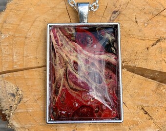 May include: Silver-toned rectangular pendant necklace with a chain. The pendant features a vibrant abstract design with red, gold, and black swirls. The pendant is set in a silver frame, creating a striking contrast. The necklace is displayed on a wooden surface.