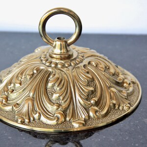 May include: Ornate brass-colored decorative lid with a ring handle. The lid features a detailed, embossed floral design. The piece is likely for a decorative container or furniture accent, adding a touch of vintage elegance.