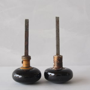 May include: Two black ceramic door knobs with brass accents and metal shafts. The knobs are round and have a slightly domed shape.