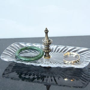 May include: A rectangular clear glass tray with a radiating sunburst pattern, featuring a central brass-toned metal post. A dark green jade-style bangle, a thin silver ring, and a gold-toned metal cuff bracelet are resting on the glass surface.
