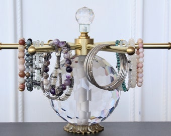 May include: A gold-toned metal jewelry display stand with a faceted crystal base and a crystal ball finial. The stand holds various bracelets in shades of pink, purple, and silver. The stand is approximately 12 inches tall.