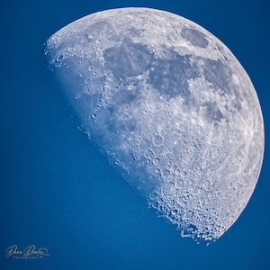 May include: A close-up of the moon in a crescent phase, showing its surface with craters and a blue sky background.