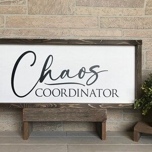 May include: A wooden sign with a white background and black lettering that reads "Chaos Coordinator". The sign is sitting on a small wooden stand.