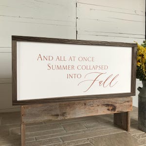 May include: A rectangular wooden sign with a dark brown frame and white background. The sign features the quote "AND ALL AT ONCE SUMMER COLLAPSED INTO Fall" in a warm brown color. The sign is displayed on a rustic wooden stand.
