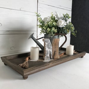 May include: A wooden tray with a decorative border and four legs. The tray is filled with a variety of items, including a metal watering can, two white candles, a wooden letter "M", and a decorative rope.