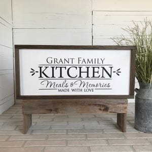 May include: A rustic wooden sign with a white background and black text. The sign reads "Grant Family Kitchen - Meals & Memories - Made with Love".