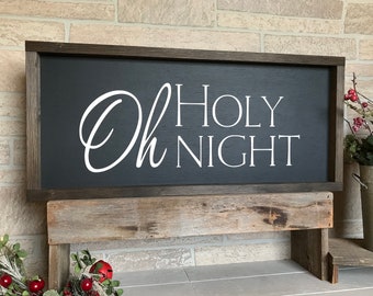 May include: A dark gray wooden sign with a white inscription that reads "Oh Holy Night". The sign is sitting on a rustic wooden stand.