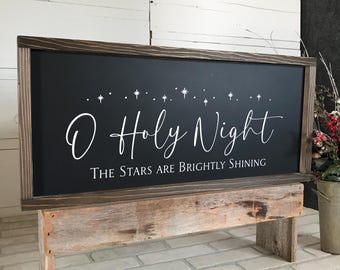 May include: A rectangular wooden sign with a dark chalkboard surface. The sign reads "O Holy Night" in elegant white script, with the phrase "The Stars Are Brightly Shining" below. The sign is framed in a dark brown wood and rests on a rustic wooden stand.