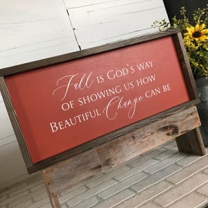 May include: A rectangular wooden sign with a dark brown frame and a red background. The sign displays the white text "Fall is God's Way of Showing Us How Beautiful Change Can Be". The sign is propped up on a rustic wooden stand.