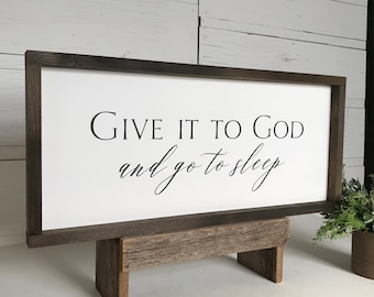 May include: A white wooden sign with a brown frame, sitting on a rustic wooden stand. The sign reads "Give it to God and go to sleep".