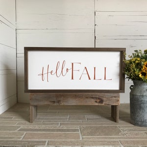 May include: A wooden sign with the text "hello FALL" in brown lettering on a white background. The sign is sitting on a rustic wooden stand.