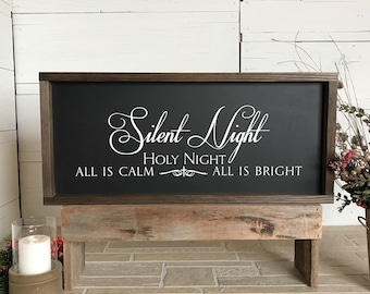 May include: A black wooden sign with white text that reads "Silent Night Holy Night All is Calm All is Bright". The sign is sitting on a wooden stand.