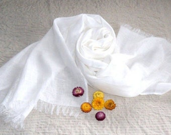 May include: A white linen scarf with a fringed edge. The scarf is folded and arranged on a textured surface with several small dried flowers.