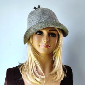 Peut inclure: Un chapeau cloche en feutre gris avec un petit nœud sur le dessus. Le chapeau a un large bord et une couronne arrondie.
