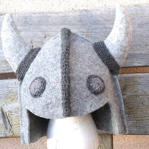 May include: A grey felt Viking helmet with horns and a grey and black knitted trim. The helmet has two grey felt circles on the front.
