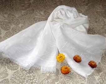 May include: A white cotton scarf with a fringed edge. The scarf is folded and laid on a patterned surface with four small yellow flowers.