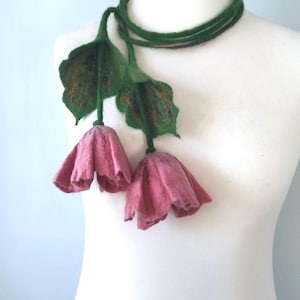 May include: A handcrafted felt necklace with two pink flower pendants, green leaves, and a green cord. The flowers have a textured finish, and the leaves have brown detailing. The necklace is shown on a white mannequin.