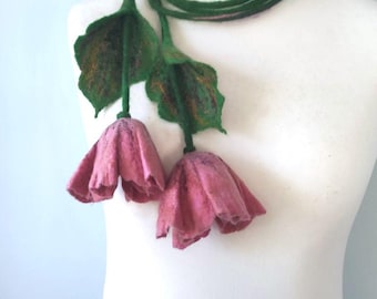 May include: A handmade felt necklace featuring two pink flower pendants with green leaves and a green cord. The flowers have a textured appearance, and the leaves have brown accents. The necklace is displayed on a white mannequin.