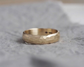 May include: A gold ring with a hammered texture. The ring is a warm, yellow color and has a slightly rounded shape. The surface of the ring has a textured, dimpled appearance. The ring is set against a soft, gray background.