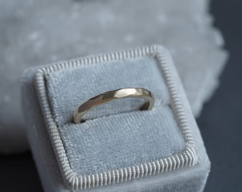 Puede incluir: Un anillo dorado con textura martillada se encuentra dentro de un joyero de terciopelo azul claro. El anillo tiene un diseño simple y elegante y una forma ligeramente curvada. El joyero tiene un borde cosido blanco.