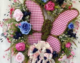May include: A pink and white gingham butterfly wreath with blue and pink flowers and a pink and white fabric bow. The wreath is decorated with greenery and has a sign that says "Wreath Whimsy by Robin".