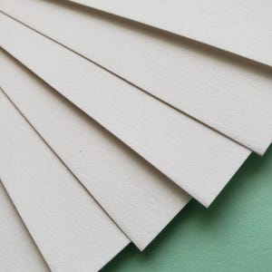 May include: A stack of off-white, rectangular paper sheets fanned out diagonally, revealing their textured surface. The paper is arranged against a soft green background, creating a contrast in color and texture. The image highlights the clean lines and subtle variations in the paper.
