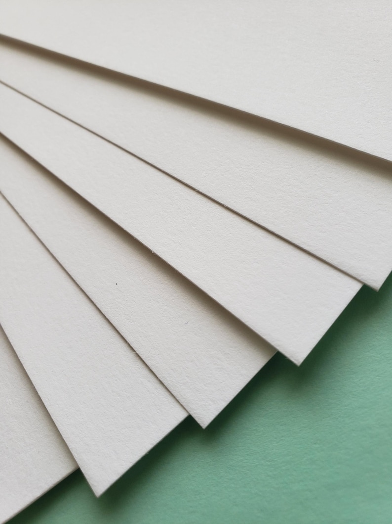 May include: A stack of off-white, rectangular paper sheets fanned out diagonally, revealing their textured surface. The paper is arranged against a soft green background, creating a contrast in color and texture. The image highlights the clean lines and subtle variations in the paper.