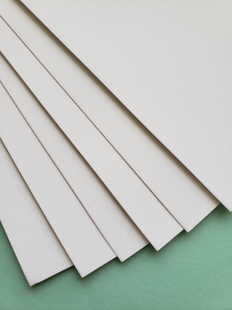 May include: A stack of off-white rectangular paper sheets, fanned out to reveal their edges. The paper has a slightly textured surface and is arranged diagonally against a green background. Ideal for art or stationery.