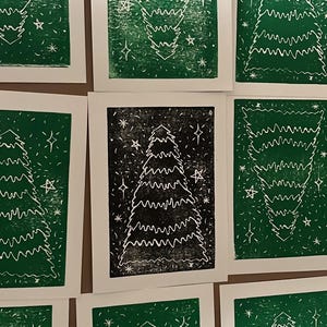 May include: A collection of handmade Christmas cards featuring a linocut design of a Christmas tree. The cards are printed in green and black ink on white cardstock, with star accents. The tree design has a hand-drawn aesthetic.