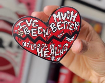 Puede incluir: Pegatina roja en forma de corazón con contorno negro y texto blanco que dice "He estado mejor pero al menos estoy sanando" con un gráfico de flor y rayo.