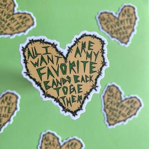 Peut inclure: Un autocollant en forme de cœur avec une bordure de fil barbelé. L'autocollant est jaune avec du texte vert qui dit "Tout ce que je veux, c'est que mes groupes préférés se réunissent".