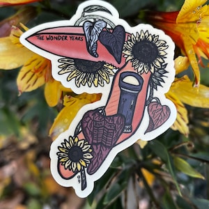 Peut inclure: Un autocollant représentant une planche de surf rouge avec le texte "The Wonder Years" et diverses illustrations florales et botaniques, notamment des tournesols, des feuilles et une lampe de poche.