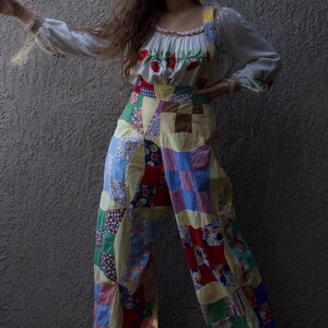 Può includere: Una tuta patchwork con una camicetta bianca. La camicetta ha maniche lunghe ed è ricamata con fiori rossi. La tuta è realizzata con varie toppe di tessuto colorato. L'outfit è uno stile vintage unico.