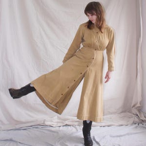 Könnte beinhalten: Ein langes, hellbraunes, geknöpftes Kleid mit einem Schlitz vorne und langen Ärmeln. Das Kleid ist aus einem leichten Stoff gefertigt und hat eine lockere Passform. Das Model trägt das Kleid mit einem Paar braunen Stiefeln.