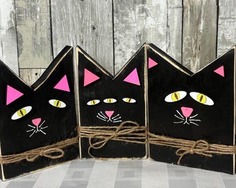 May include: Three black wooden cat decorations with pink triangle ears, yellow and white eyes, and pink noses. Each cat is wrapped with twine. The background is a weathered wood panel.