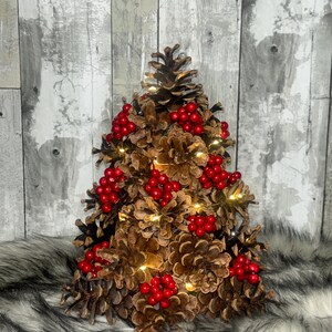 May include: A decorative Christmas tree made of pine cones, adorned with red berries and small, warm white lights. The tree is set against a gray and white wooden background and rests on a faux fur surface.