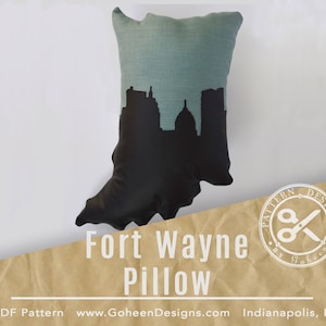 May include: A teal blue pillow with a black silhouette of the Fort Wayne, Indiana skyline. The pillow is on a brown background with the text "Fort Wayne Pillow" and "PDF Pattern www.GoheenDesigns.com Indianapolis, IN".