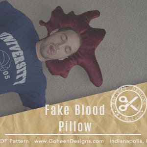 Puede incluir: Una almohada roja con forma de salpicadura de sangre con una persona acostada encima. La almohada tiene forma de salpicadura de sangre y está hecha de un material suave y mullido. La persona lleva una camisa azul con la palabra "UNIVERSITY" en ella. La almohada es una forma divertida y única de añadir un toque de humor a la decoración de tu hogar.